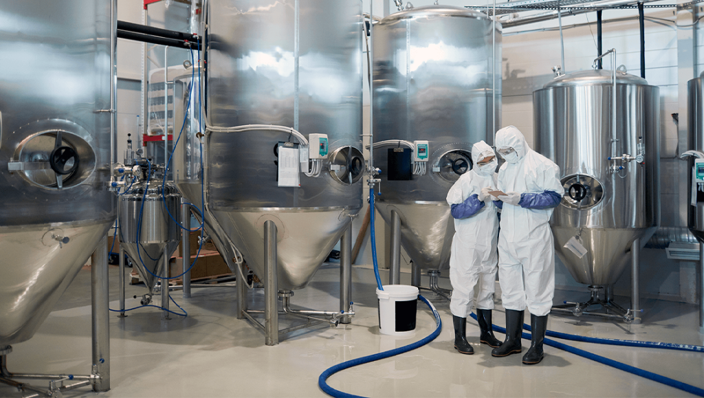 Two workers in hazmat suits inspect large stainless steel tanks in a supply chain facility.