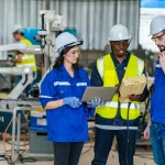 Engineers in hard hats review data on a laptop and device in a factory