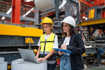 Two women in hard hats review data on a laptop in a factory setting, discussing manufacturing analytics.