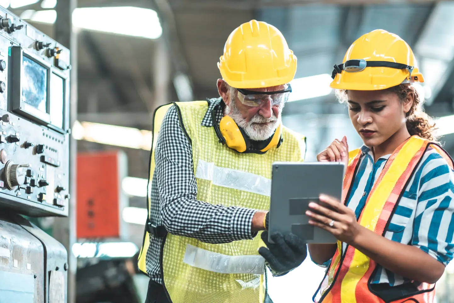 Engineers in hard hats review manufacturing software on a tablet in a factory.